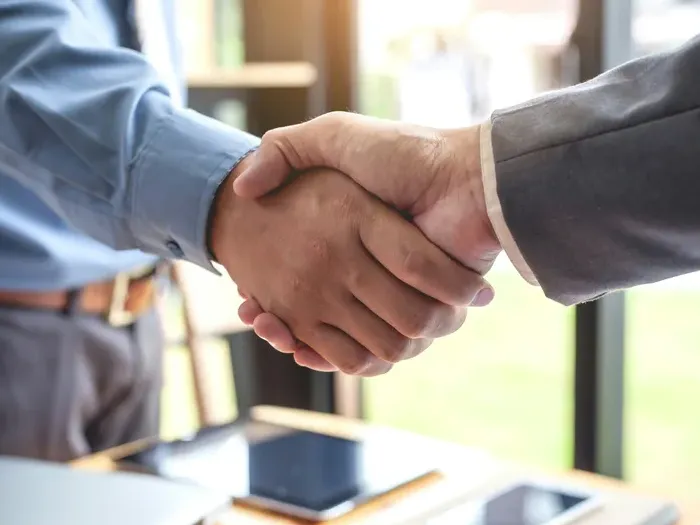 Closeup of two businessmen shaking hands at office table with digital devices in background symbolizing partnership